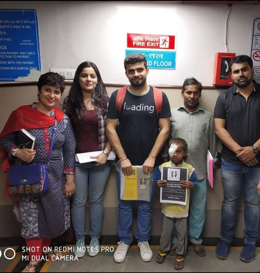 A photo of a group of sarcoma patients and advocates posing in the hallway of a hospital in India. The group includes two women, three men, and a young boy with a bandage over his eye. The group is distributing information sheets to chemotherapy patients to offer advice and support.