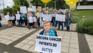A group of protesters standing on a sidewalk with a grassy area behind them, holding signs advocating for sarcoma patients, appealing to the Irish Health Service Executive and the Minister of Health. One woman stands in the foreground holding a sign that says “Sarcoma Cancer Patients Do Matter.” Several of the protesters in the background hold bundles of yellow balloons symbolising sarcoma patients.