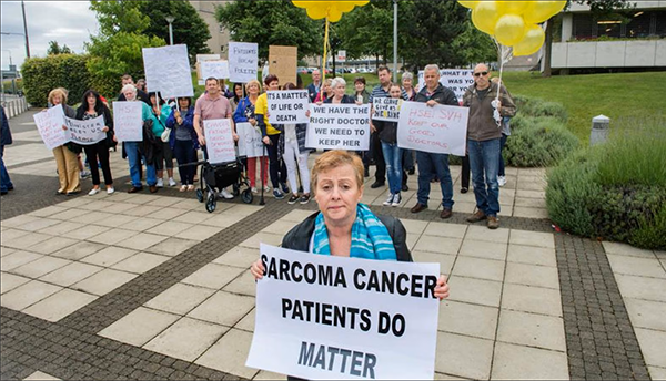 A group of protesters standing on a sidewalk with a grassy area behind them, holding signs advocating for sarcoma patients, appealing to the Irish Health Service Executive and the Minister of Health. One woman stands in the foreground holding a sign that says “Sarcoma Cancer Patients Do Matter.” Several of the protesters in the background hold bundles of yellow balloons symbolising sarcoma patients.