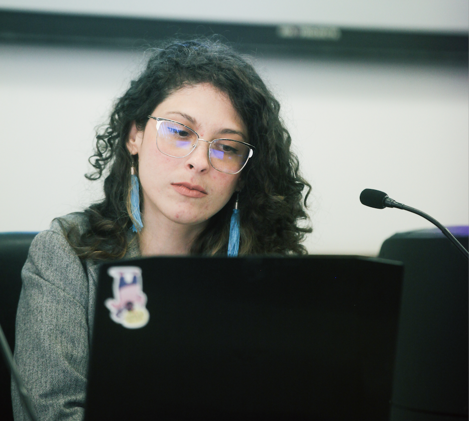 Woman ? one of the two in the cover photo (Carolina) - sitting at a desk and looking at a laptop screen, in front of a microphone, pensive.