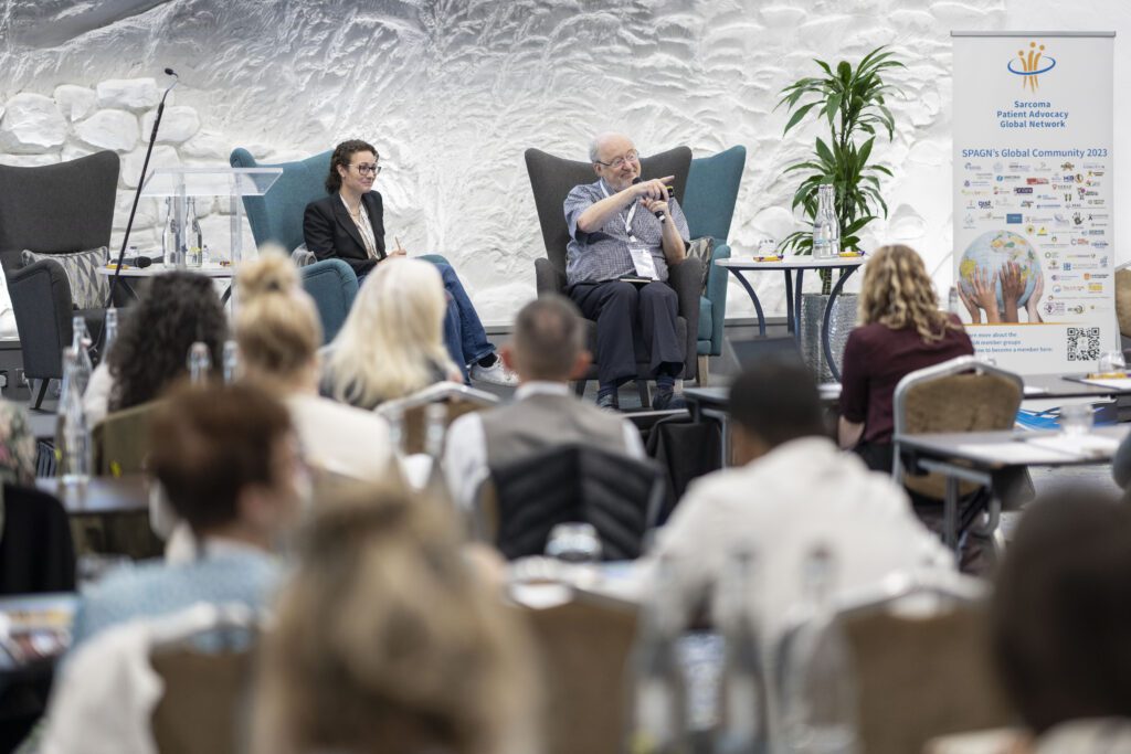 A photo of two people seated on a stage at a small conference. The man is pointing to someone in the audience.