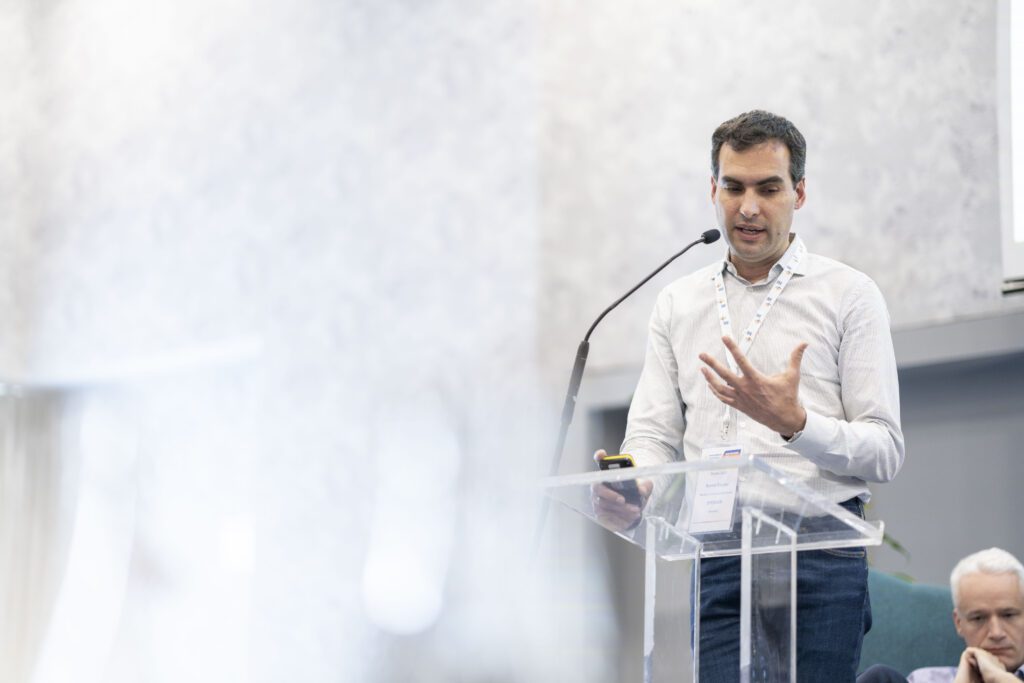 A photo of a man standing at a clear plexiglass lectern speaking at a conference.