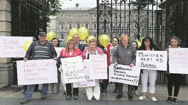 A group of protesters standing in front of an ornate iron gate with a large grey building in the background. They are holding signs advocating for sarcoma patient needs and appealing to the Minister of Health and the Health Service Executive. Many are also holding yellow balloons symbolising sarcoma patients.