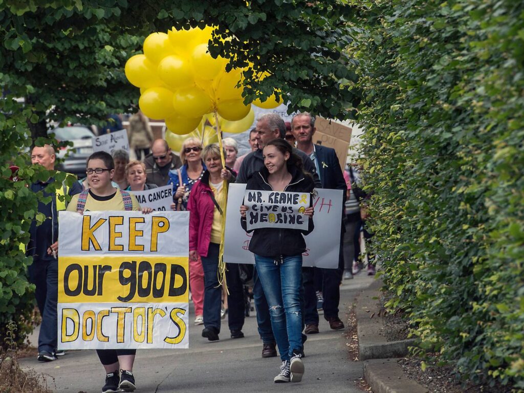 A group of protesters walking as a group down a sidewalk with trees and bushes around and overhead. At the front of the group are two young people carrying signs. On the left, a young person carries a large sign almost as tall as they are which reads “Keep our good doctors” in yellow and black handwritten letters. On the right, a young person carries a smaller sign held at chest height that reads “Mr. Keane give us a hearing” in yellow and black handwritten letters, with a yellow ribbon drawn to symbolise sarcoma patients. The other protesters in the background are also carrying yellow balloons.